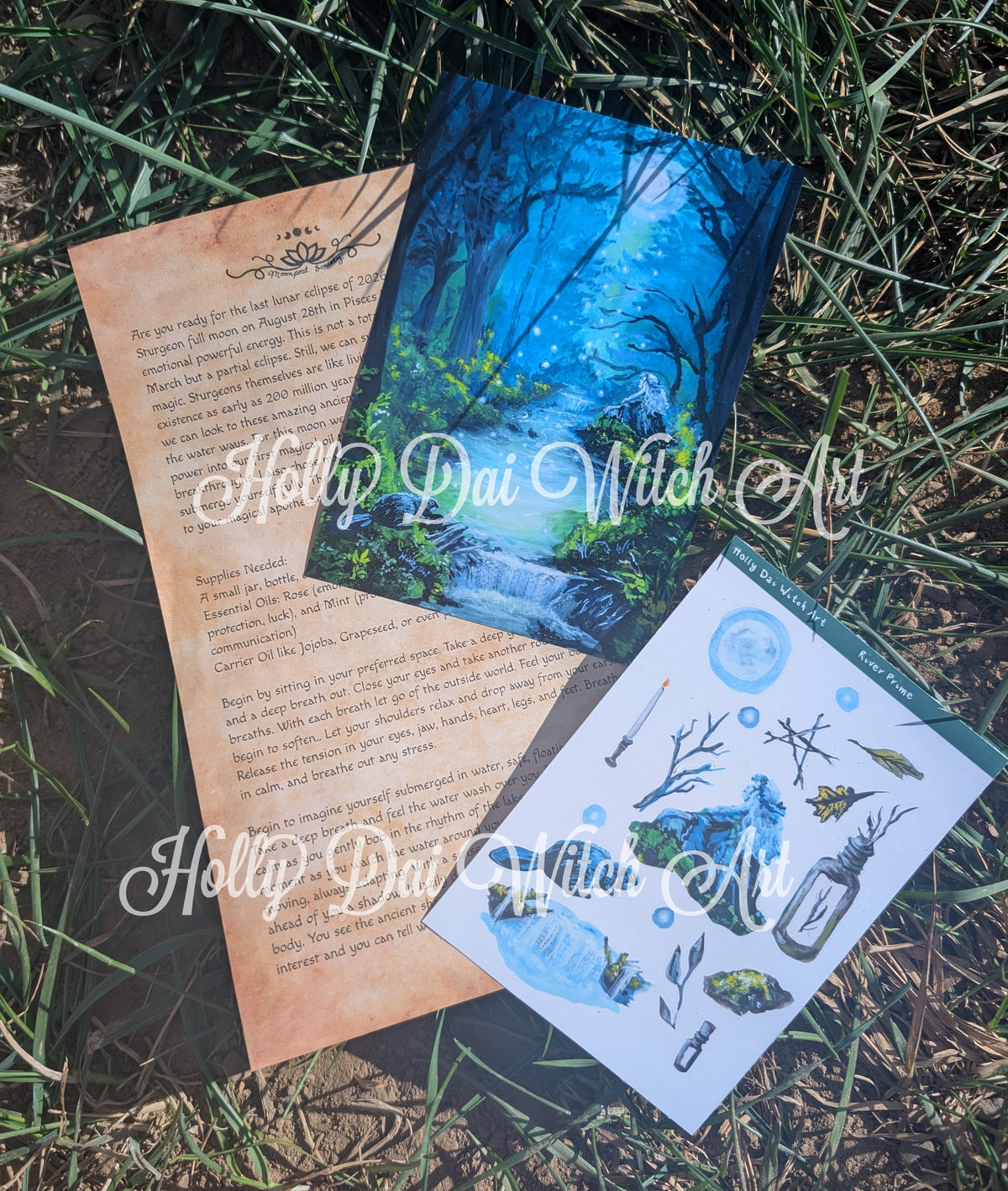 Three art pieces of paper from Holly Dai Witch Art are arranged on a grassy outdoor surface. The set includes a parchment-style ritual, a blue forest illustration print, and a white sticker sheet with blue watercolor illustrations of a witch, potion bottles, and a waterfall.