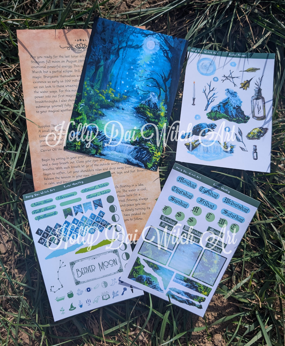 A ritual, three sheets of decorative planner stickers and a printed card featuring a blue forest stream scene, arranged on grass. The sheets include illustrated stars, moons, leaves, waterfalls, and text boxes.
